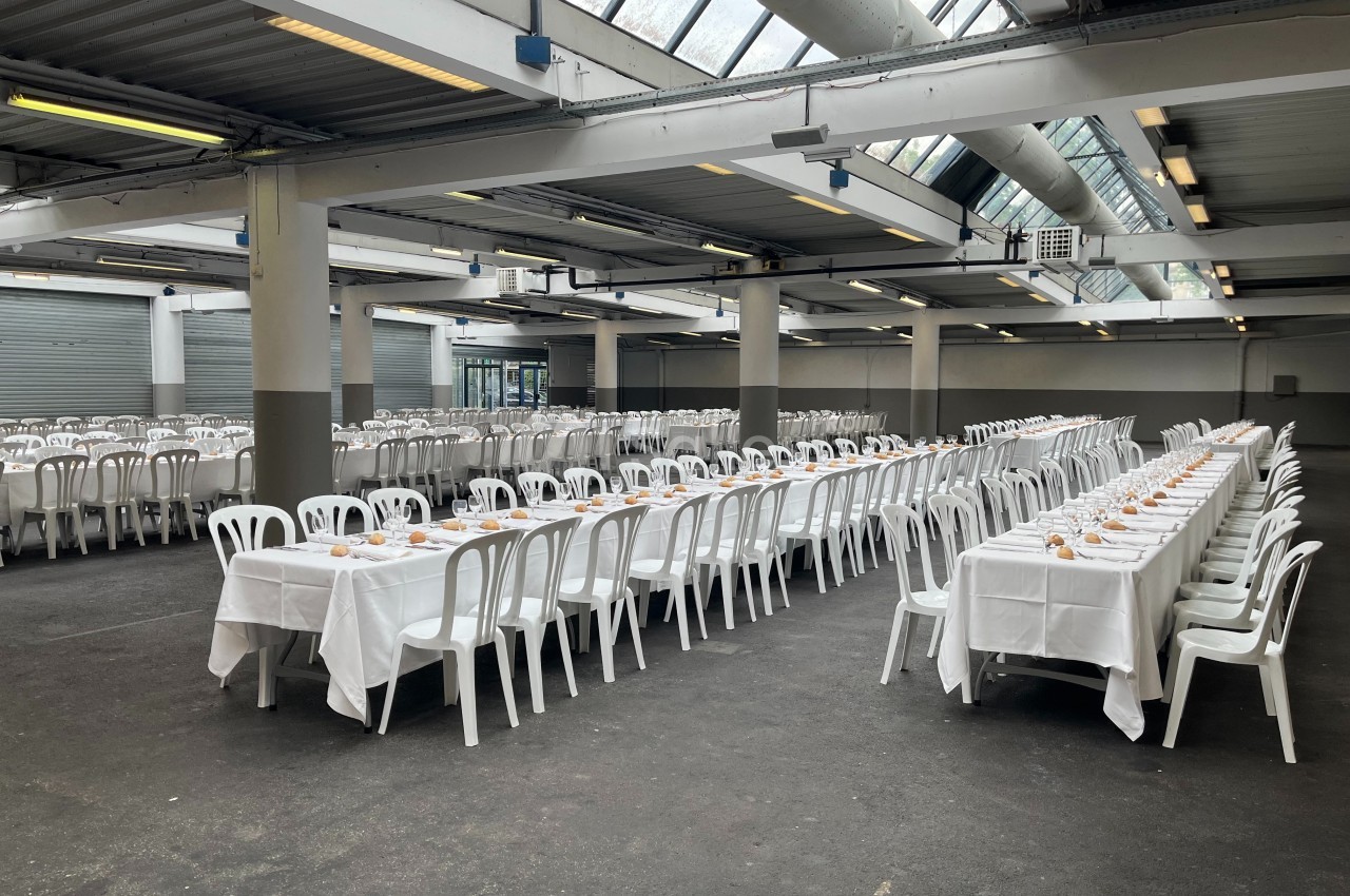 Grande salle avec de longues tables dressées de nappes blanches et alignées, entourées de chaises blanches en plastique.