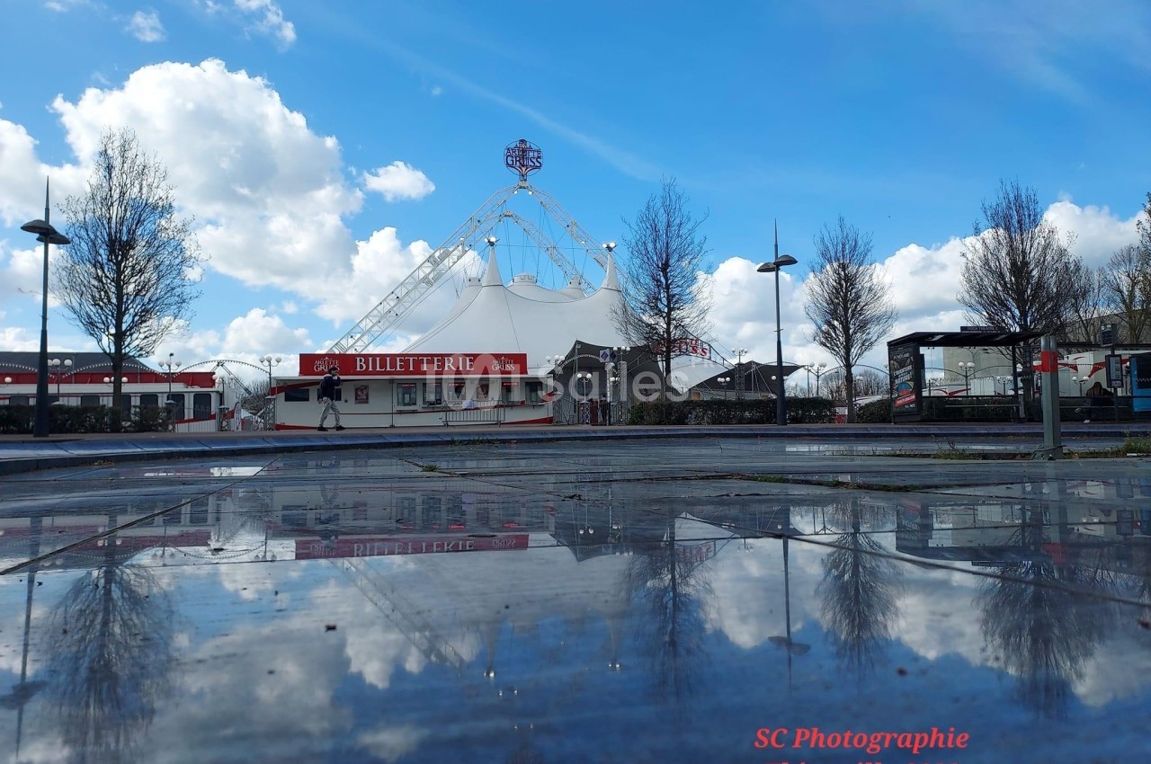Reflet d'un chapiteau blanc et d'une billetterie rouge sur une surface mouillée, sous un ciel nuageux.