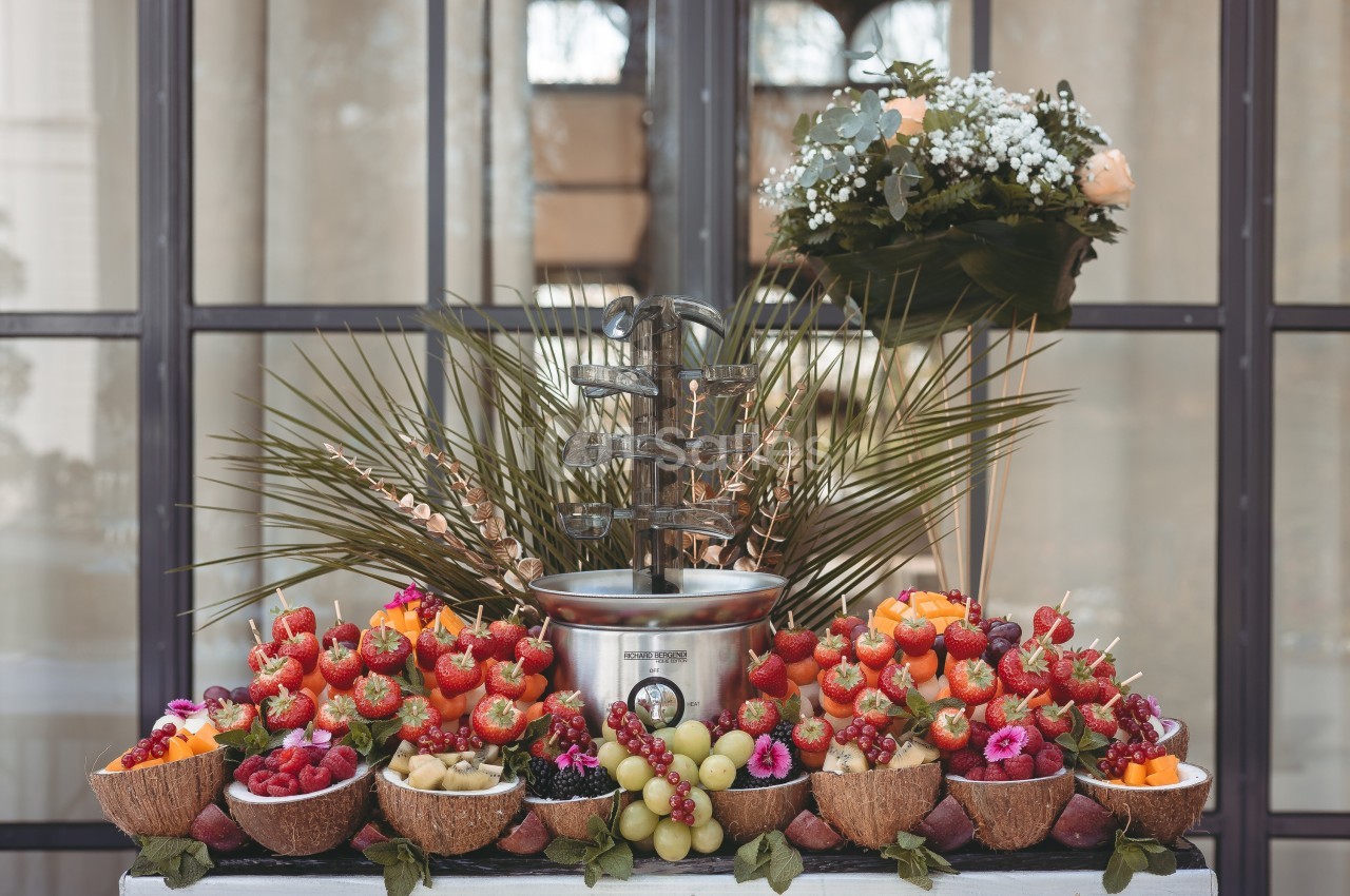 Fontaine à chocolat entourée de fruits frais variés, disposés sur une table décorée de feuilles et de fleurs.