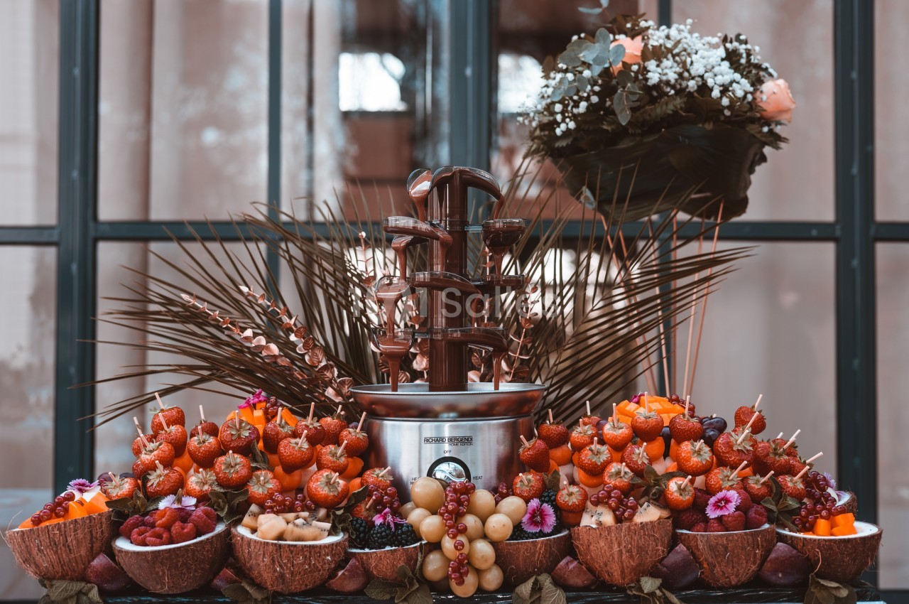 Fontaine de chocolat entourée de fruits frais, brochettes et décorations florales sur une table élégante.