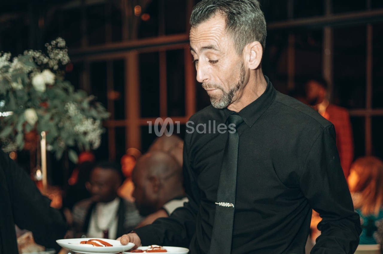 Un serveur en chemise noire tenant deux assiettes garnies, dans une salle éclairée avec des invités en arrière-plan.