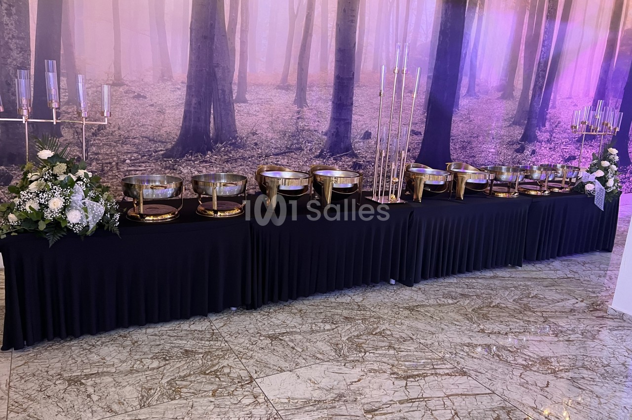 Table drapée de noir avec des plats en métal doré, entourée de chandeliers et de compositions florales, devant un décor…