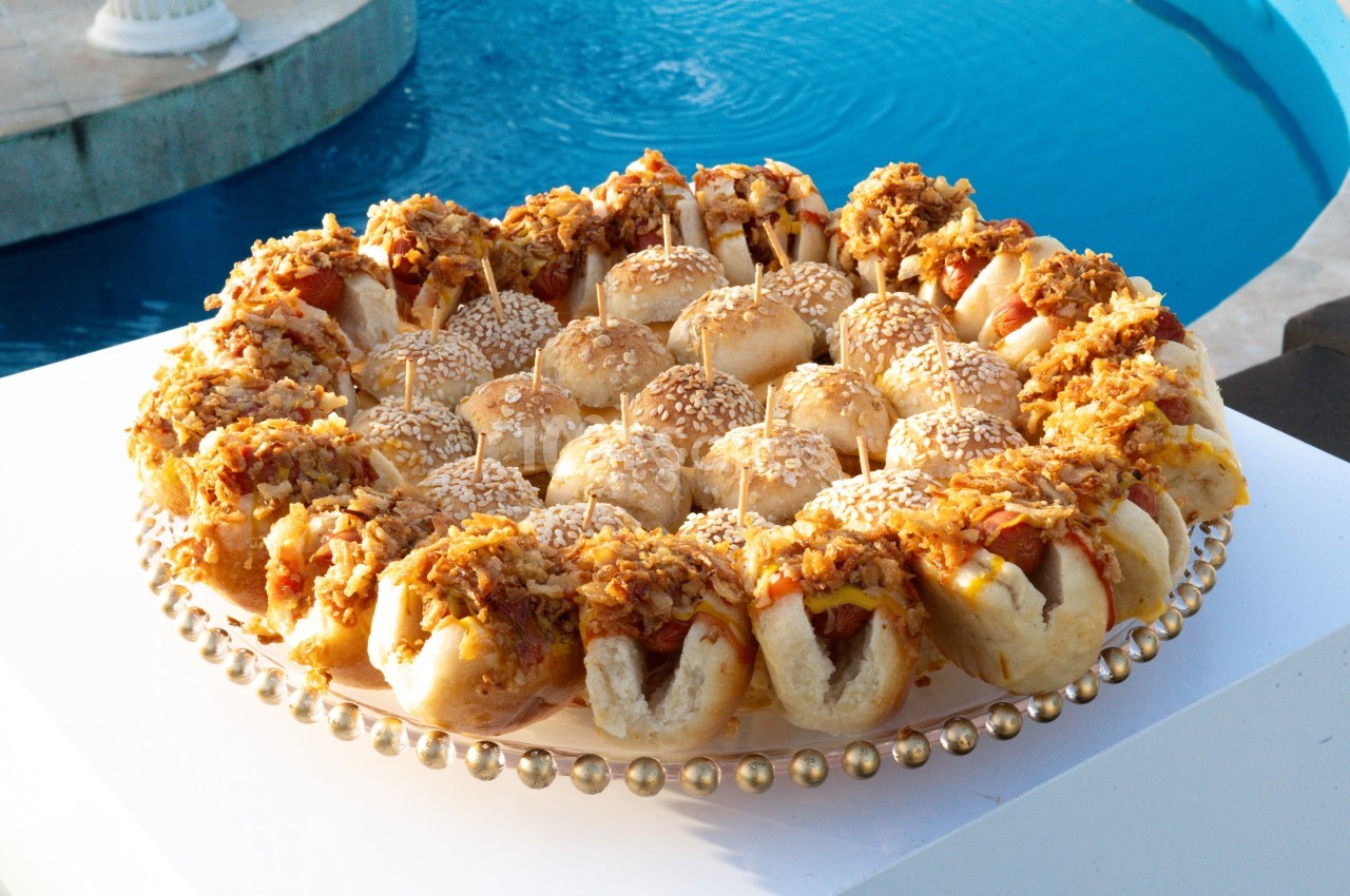 Plateau circulaire de petits pains garnis de saucisses, fromage et oignons, présenté près d'une piscine.