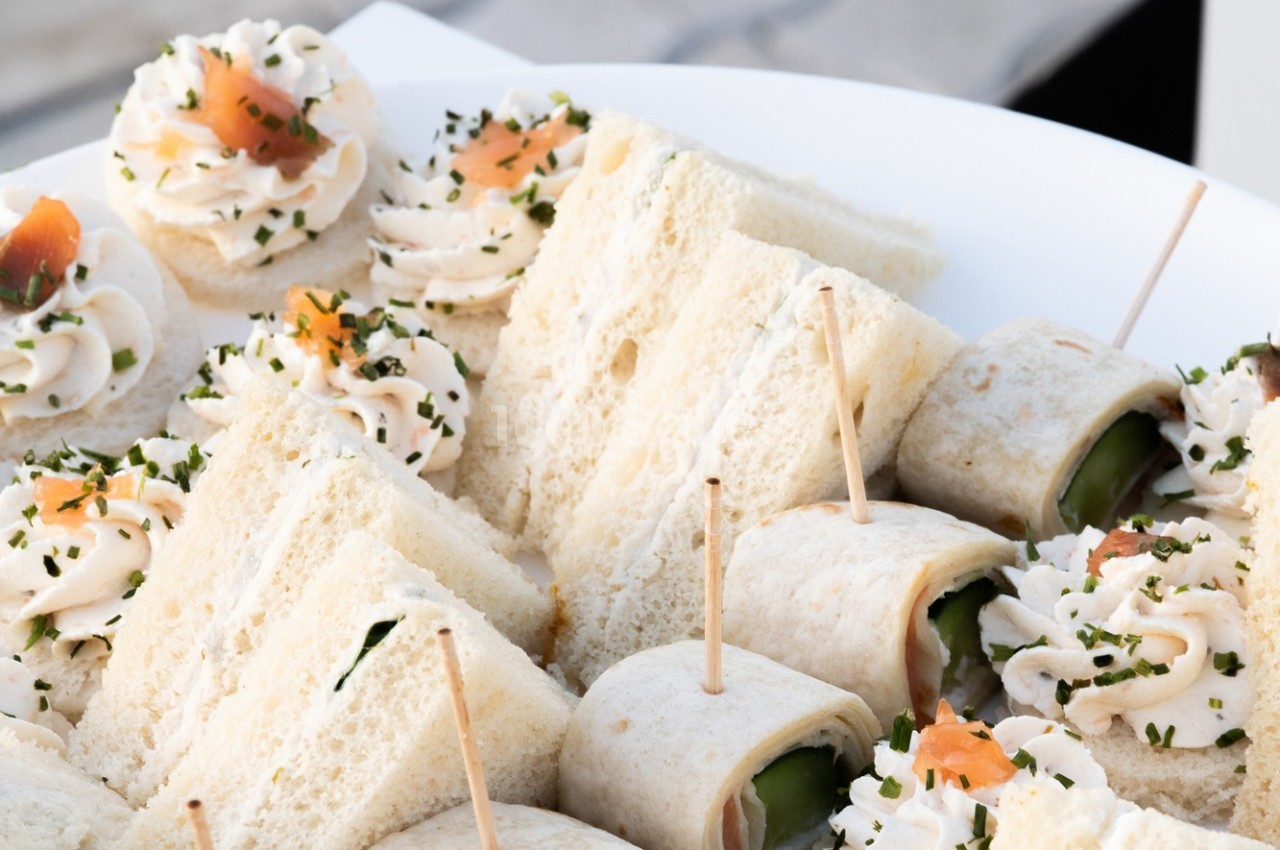 Assortiment de sandwichs, roulés et canapés garnis de saumon, crème et herbes, présentés sur un plateau blanc.
