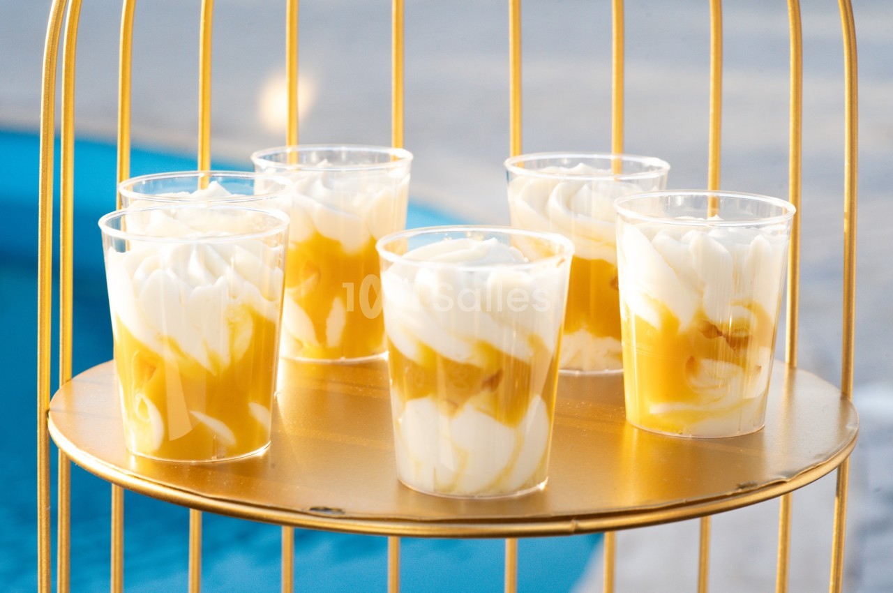 Cinq verrines de dessert à la crème et au coulis jaune, présentées sur un plateau doré près d'une piscine.