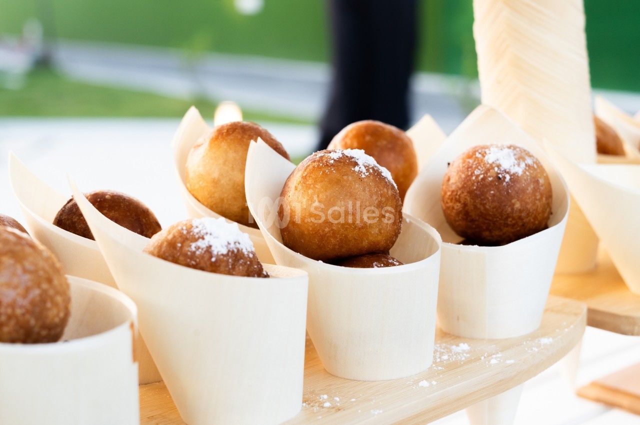 Beignets dorés saupoudrés de sucre, présentés dans des cônes en bois sur une table lumineuse.