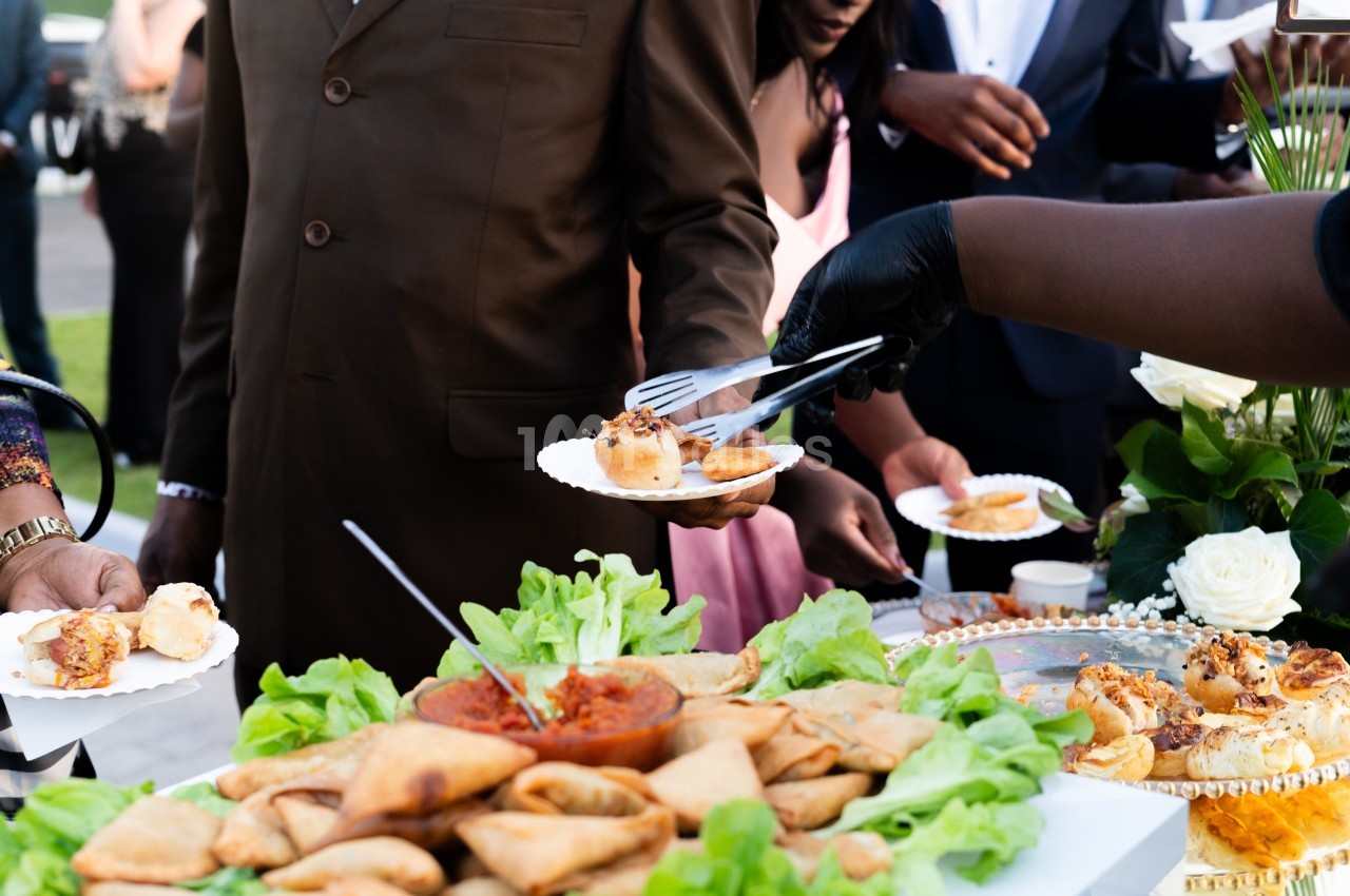 Des invités se servent des amuse-bouches sur un buffet garni de samoussas, salade et sauce, lors d'un événement en extérieur.