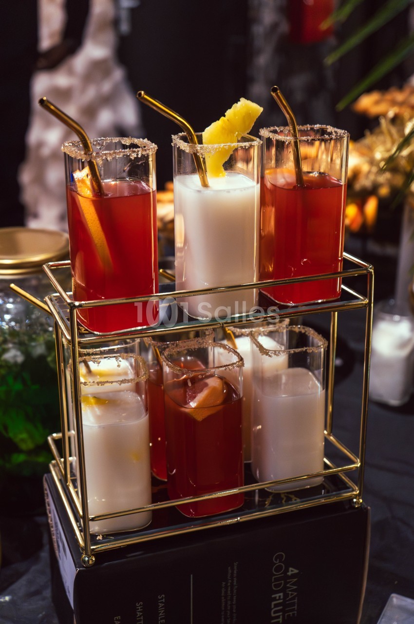 Six verres de cocktails variés avec garnitures, posés sur un support doré à deux étages.