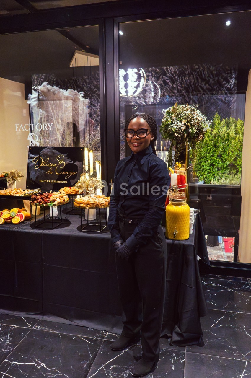 Une personne souriante en tenue professionnelle devant une table de buffet garnie de plats et de boissons.