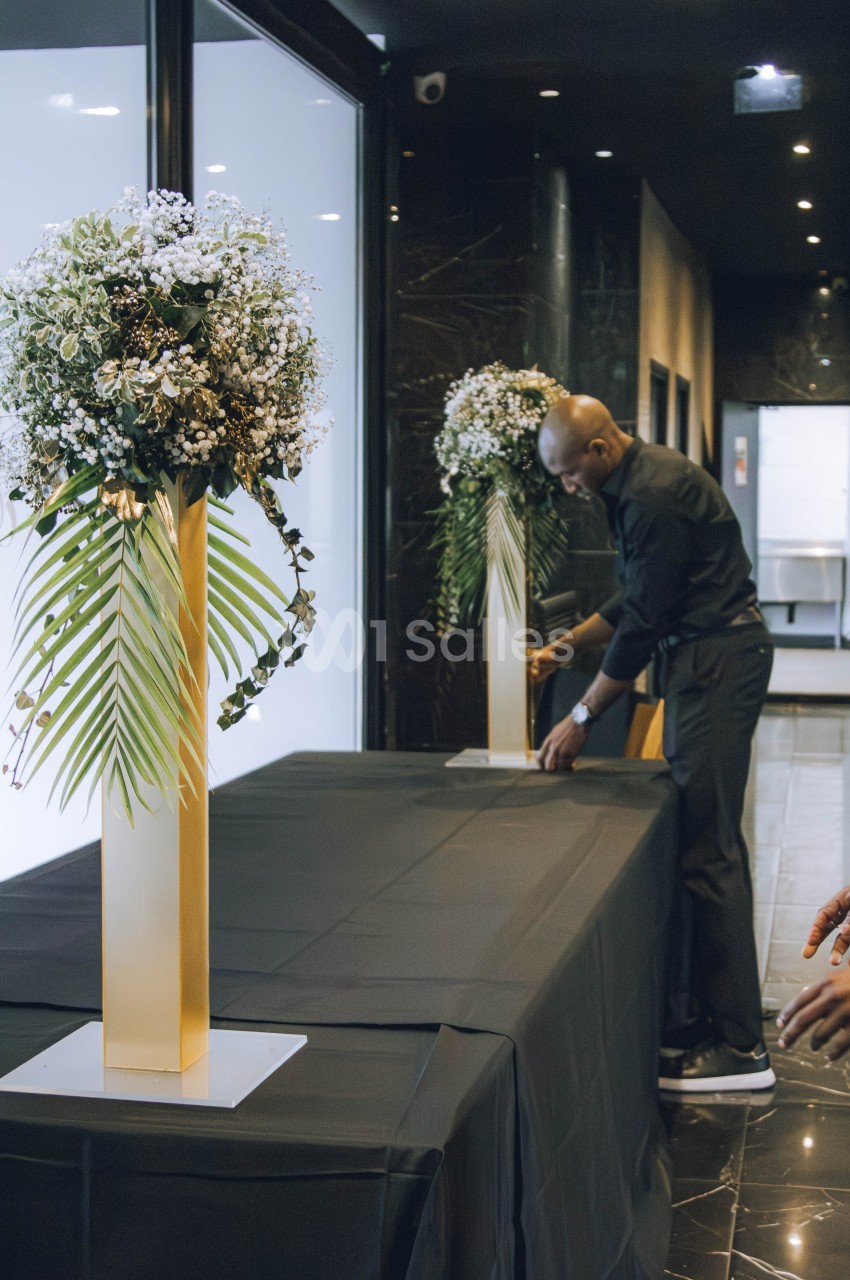 Un homme installe une table noire décorée de grands bouquets sur des supports dorés dans un espace intérieur élégant.