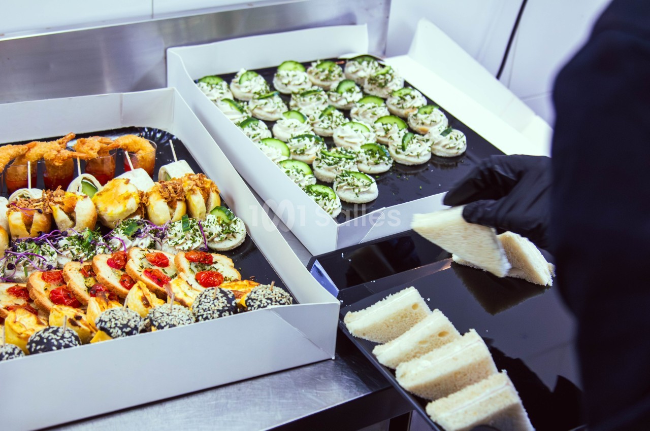 Plateaux de petits fours salés et sandwiches préparés dans une cuisine, avec une personne en gants noirs.