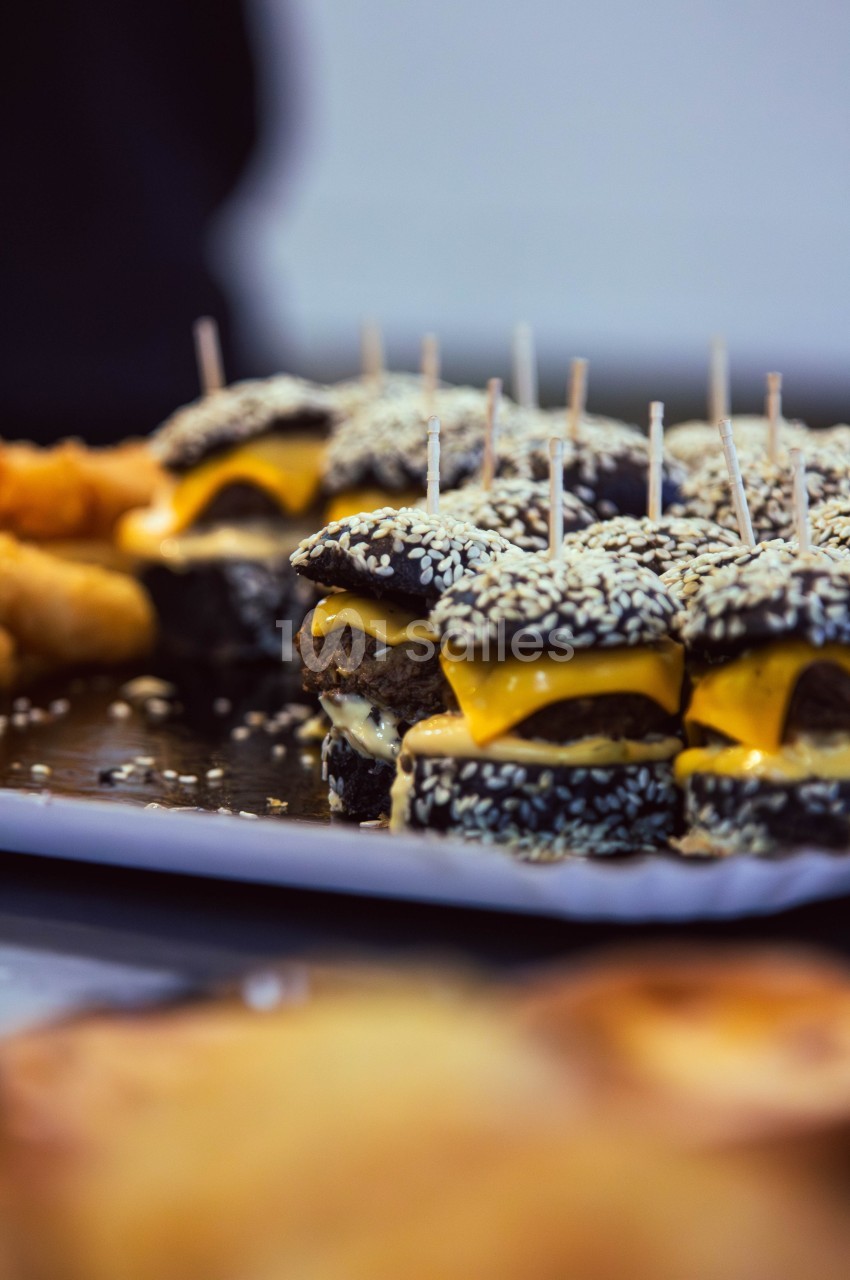 Mini-burgers au pain noir et graines de sésame, garnis de fromage et servis sur un plateau avec des amuse-bouches.