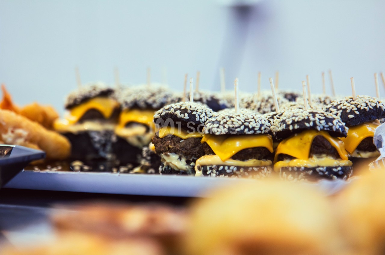 Mini-burgers au pain noir garnis de fromage fondu et de graines de sésame, alignés sur un plateau.