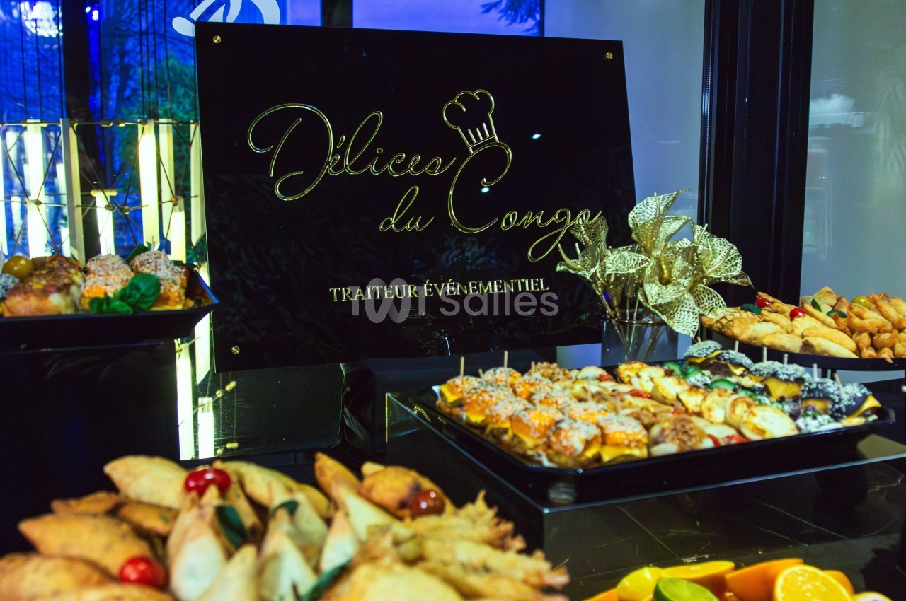 Plateau de pâtisseries et amuse-bouches variés disposés devant un panneau ’Délices du Congo, traiteur événementiel’.