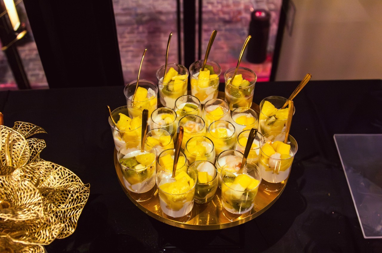 Verrines de fruits frais, principalement mangue et kiwi, présentées sur un plateau doré avec des cuillères dorées.