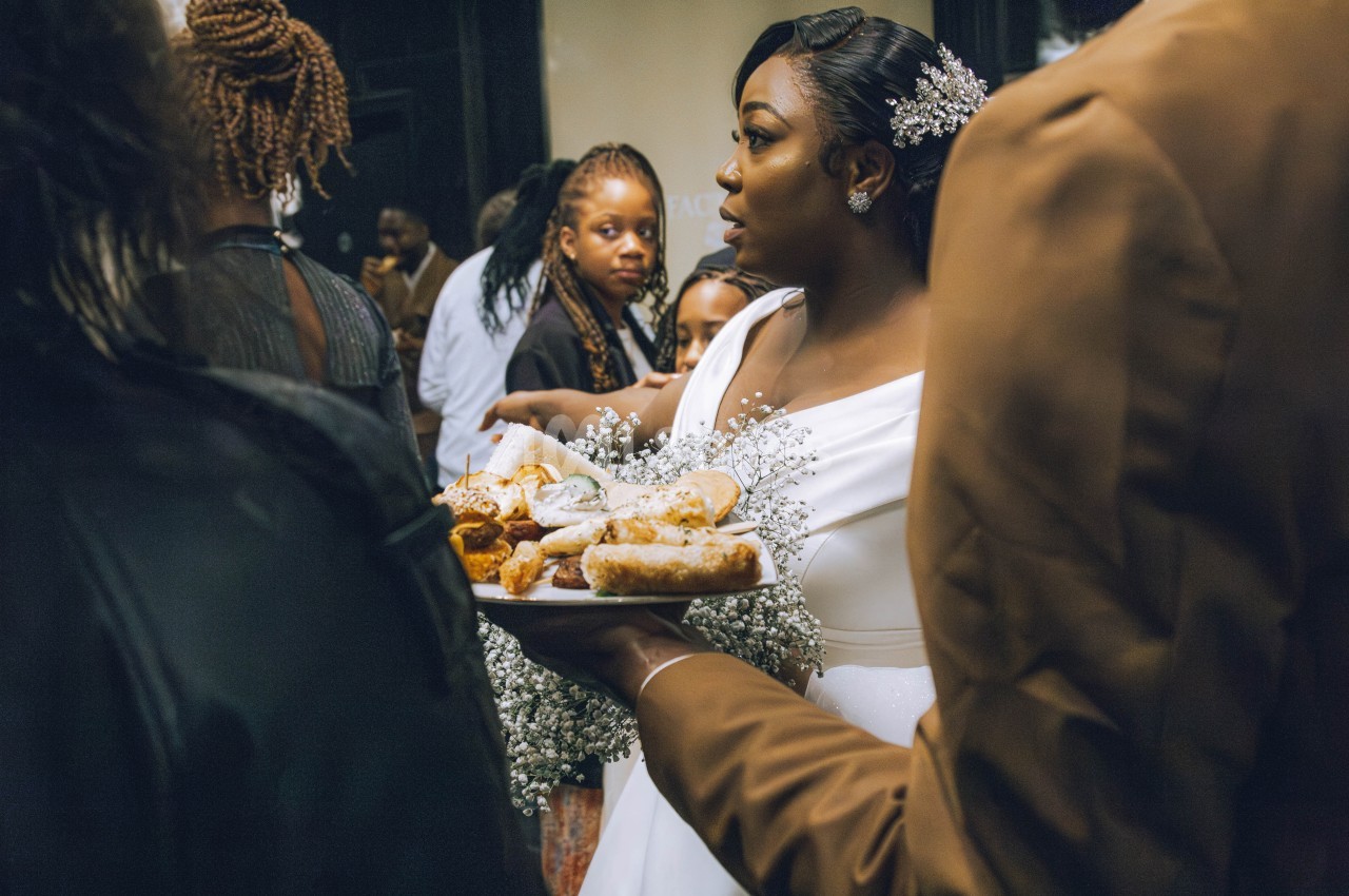 Une mariée en robe blanche tient un plateau de pâtisseries entourée d'invités lors d'une réception.