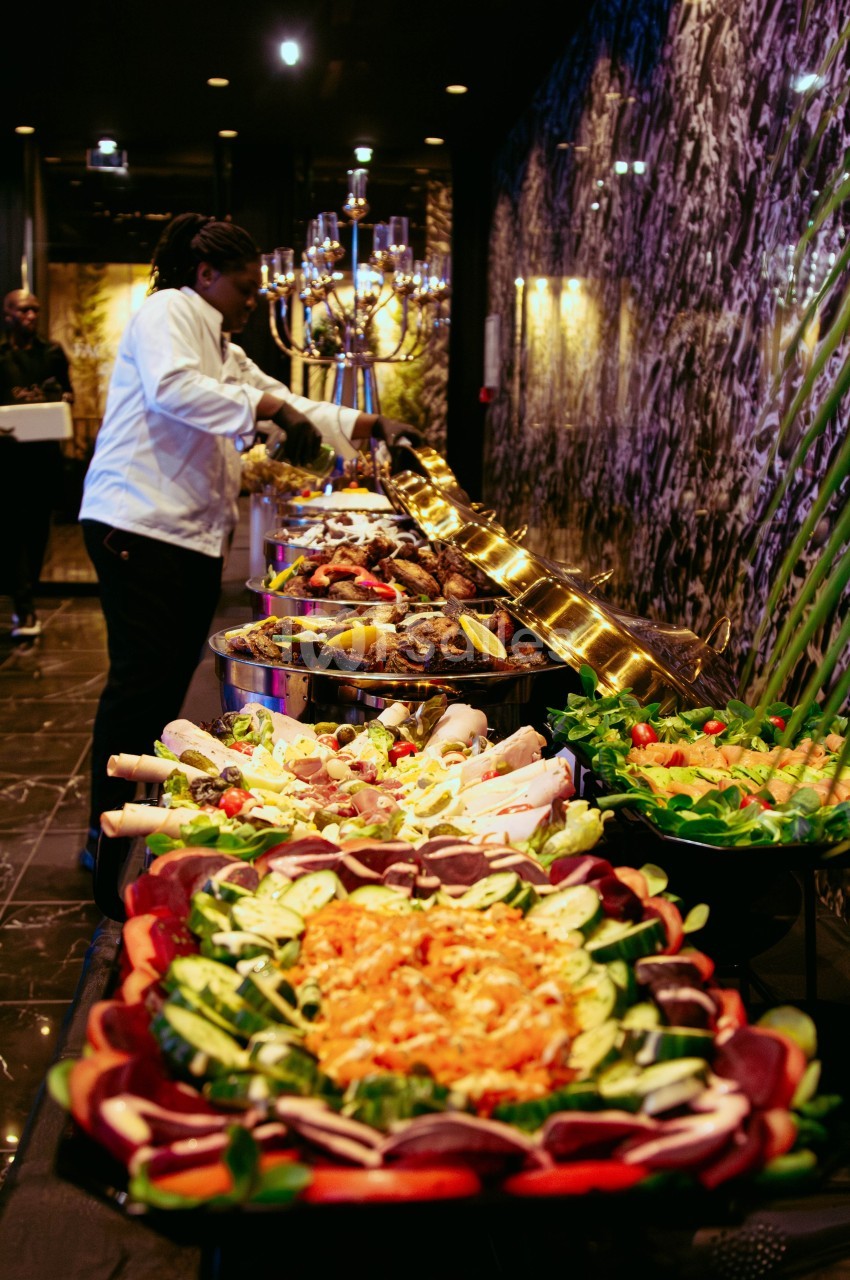 Buffet varié avec plats colorés, légumes frais, viandes grillées et chef en train de préparer les assiettes.