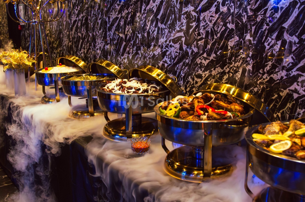 Buffet avec plats variés présentés dans des récipients en métal, entourés de vapeur pour un effet visuel.