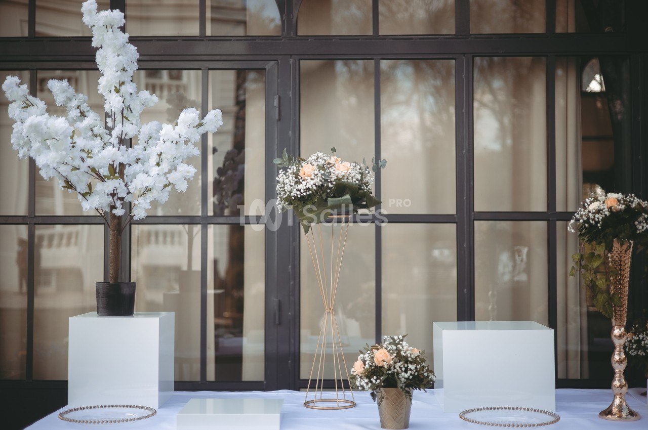 Décoration florale composée de bouquets et d'arbres blancs sur des socles devant une grande baie vitrée.
