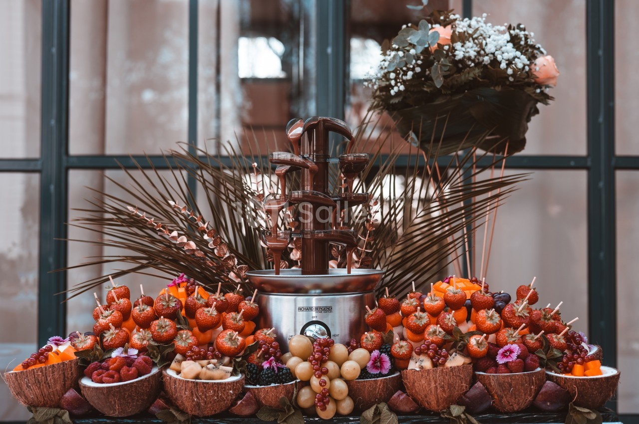 Fontaine à chocolat entourée de fruits frais, de fleurs et de décorations végétales, disposés sur une table.