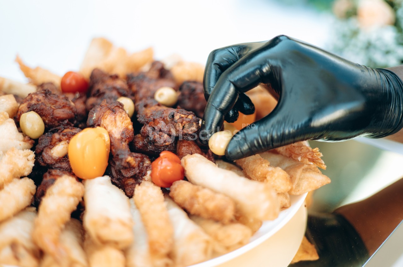Main gantée noire prenant une olive sur un plateau de bouchées variées, incluant légumes et aliments frits.