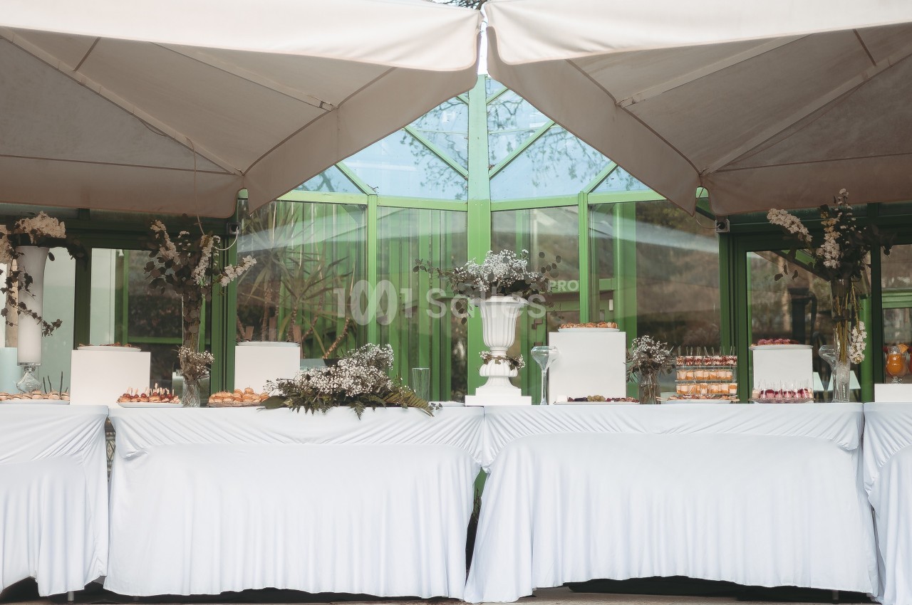 Tables décorées avec nappes blanches, ornées de fleurs et de desserts, disposées sous des parasols près d'une verrière.