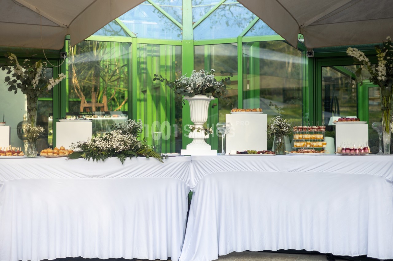 Buffet décoré avec nappes blanches, desserts variés et compositions florales devant une verrière verte.