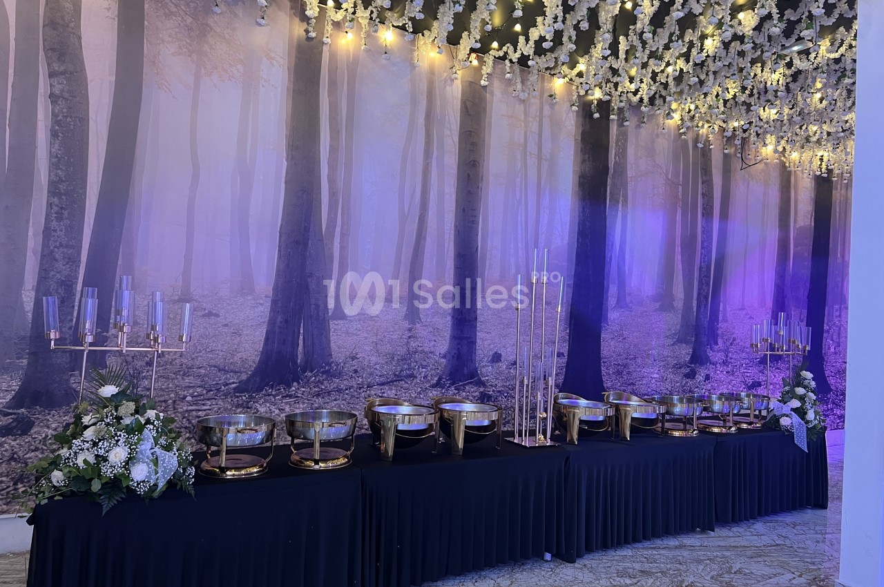 Buffet élégant avec plats en métal doré sur une table noire, décoré de fleurs blanches et éclairé par des guirlandes…