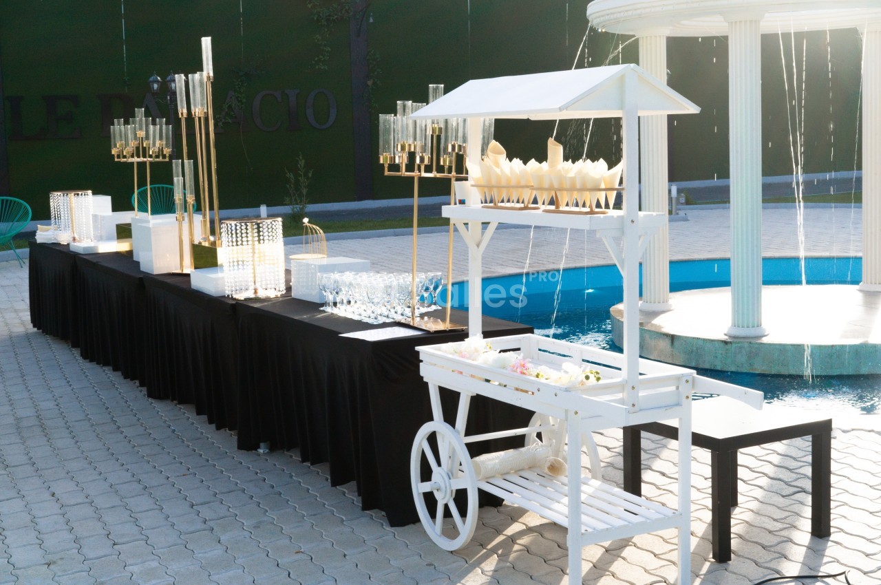 Stand de buffet en extérieur avec une table noire garnie de verres et décorations, près d'une fontaine blanche.