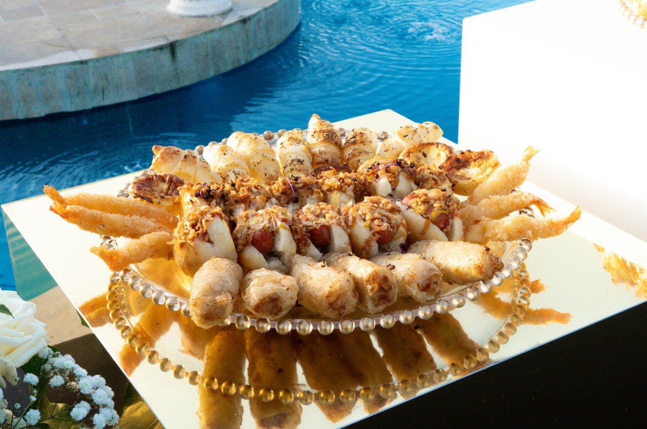 Plateau doré avec assortiment de bouchées apéritives, incluant des rouleaux frits et des crevettes, près d'une piscine.