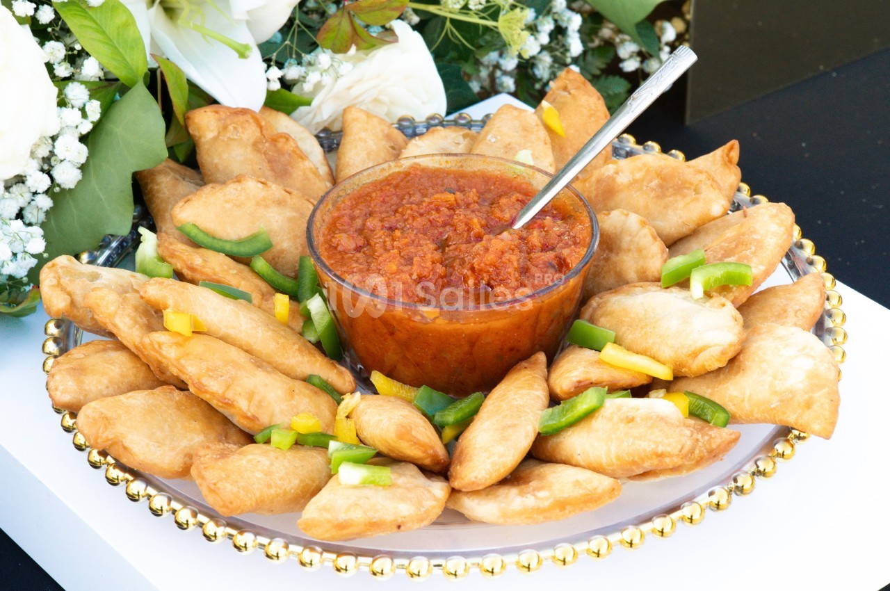 Assiette de samoussas dorés entourant un bol de sauce rouge, décorée de morceaux de poivrons colorés.