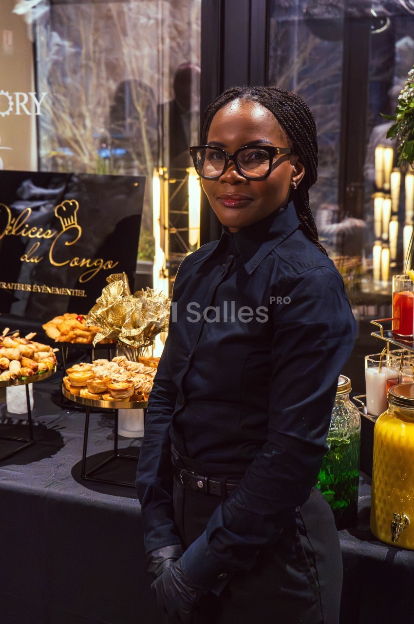 Une femme en tenue professionnelle devant une table garnie de plats et de boissons lors d'un événement.