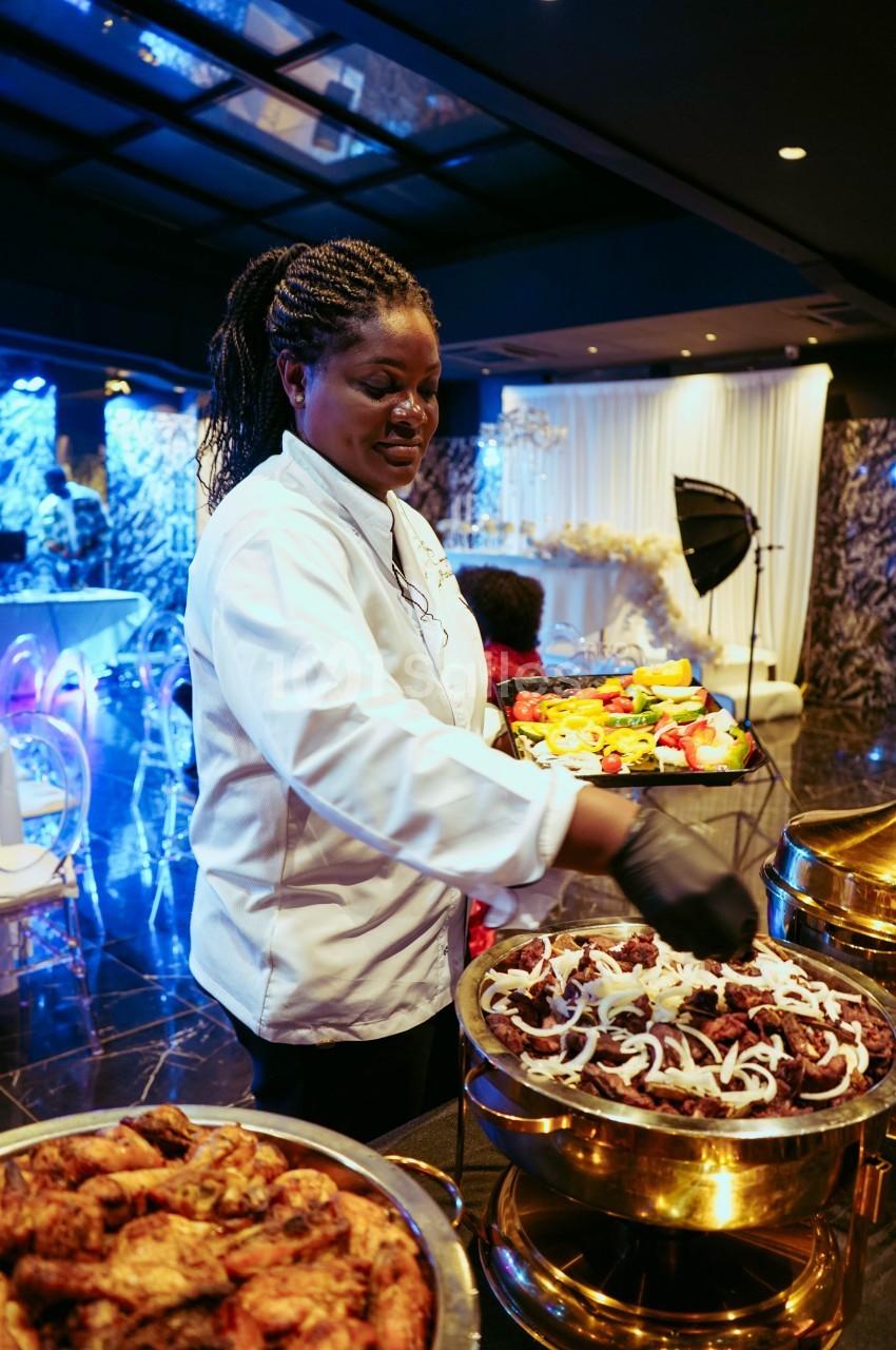 Une cheffe en tenue blanche dispose des plats garnis de viande et légumes dans une salle de réception élégante.