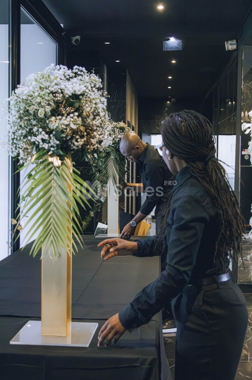 Deux personnes en tenue noire arrangent une table décorée de grands bouquets floraux dans un espace intérieur sombre.