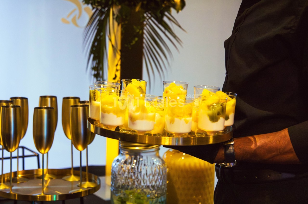 Plateau de verrines garnies de fruits jaunes et de crème, tenu par une personne vêtue de noir lors d'un événement.