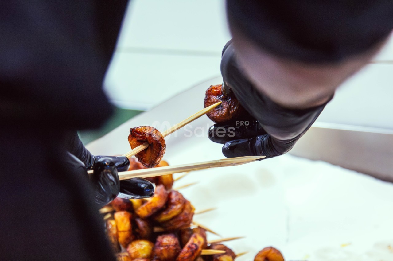Deux mains gantées enfilent des morceaux de banane plantain caramélisés sur des brochettes en bois.
