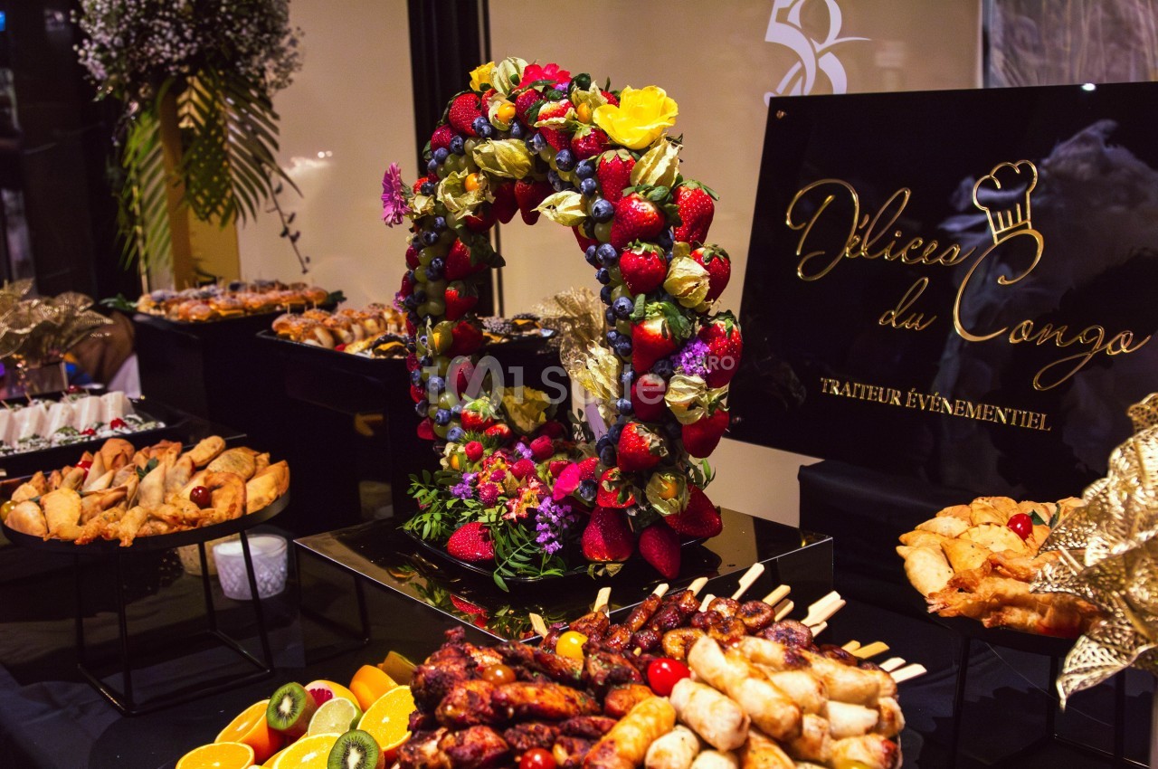 Buffet coloré avec brochettes, fruits frais, pâtisseries et une couronne décorative de fruits devant un panneau traiteur.
