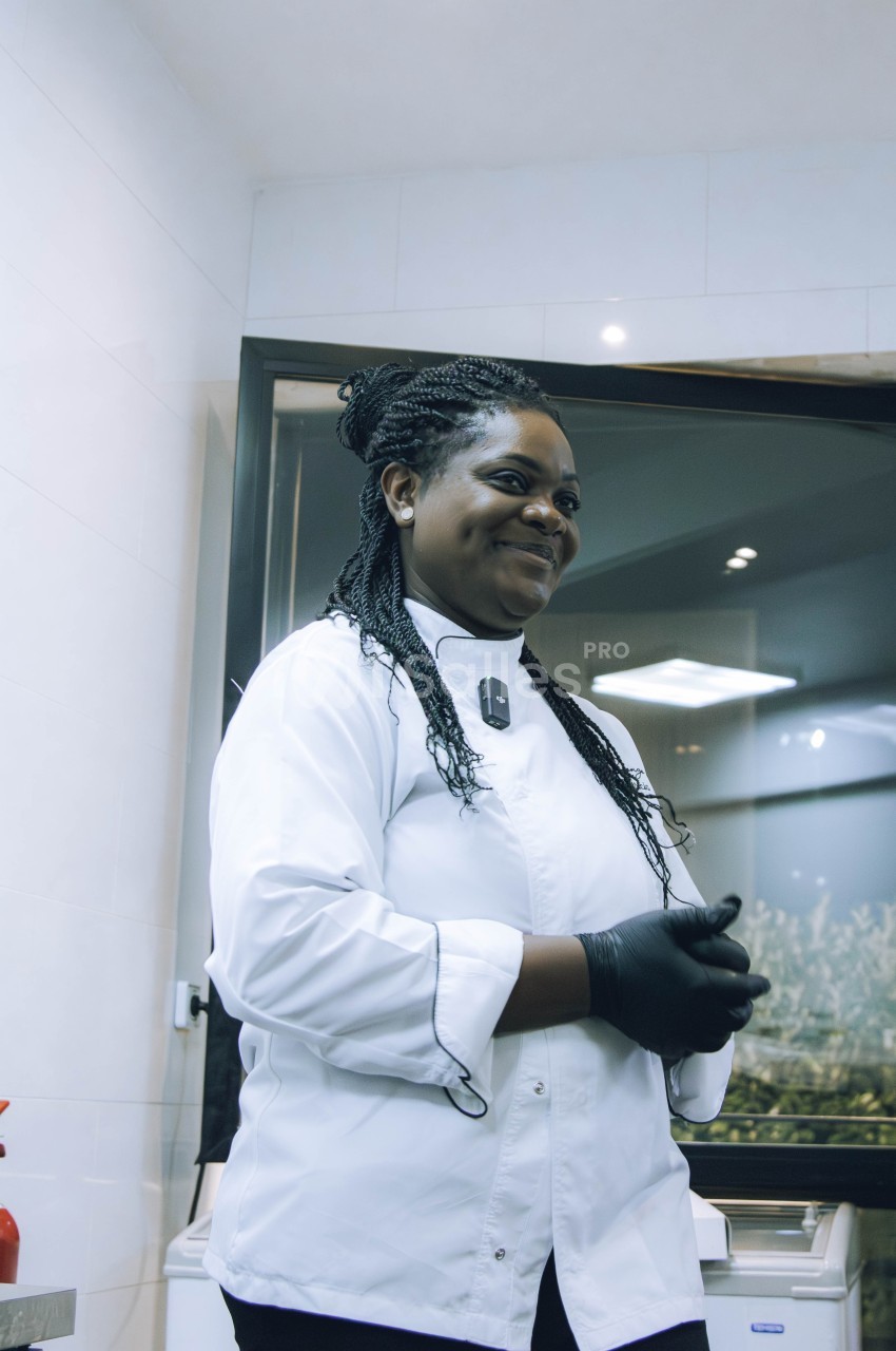 Une cheffe en blouse blanche et gants noirs se tient debout dans une cuisine moderne, souriante et détendue.