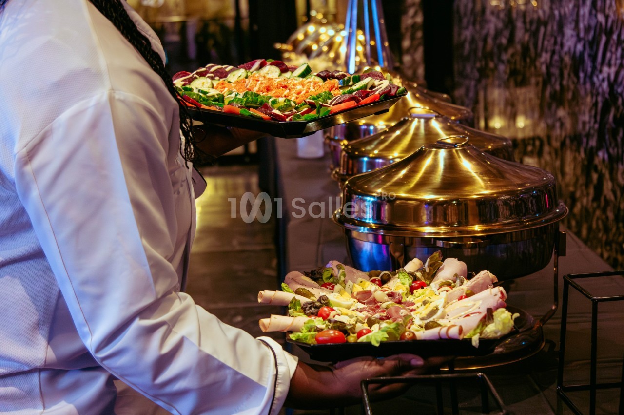 Un serveur portant des plateaux de charcuterie et de légumes devant un buffet avec des plats en métal doré.