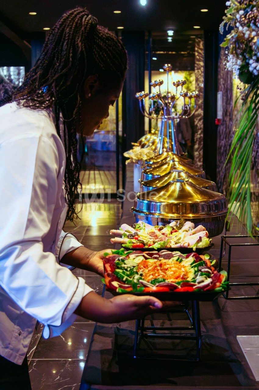 Une cheffe dispose un plat de salade sur une table de buffet garnie de mets variés dans un cadre élégant.