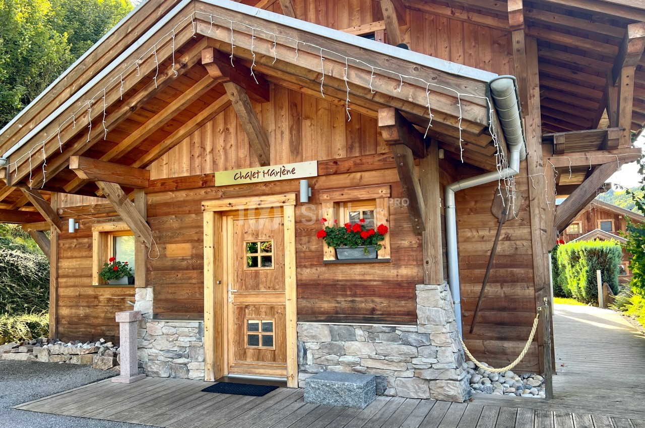 Chalet en bois avec balcon, jardinières fleuries et toit incliné, entouré de végétation.