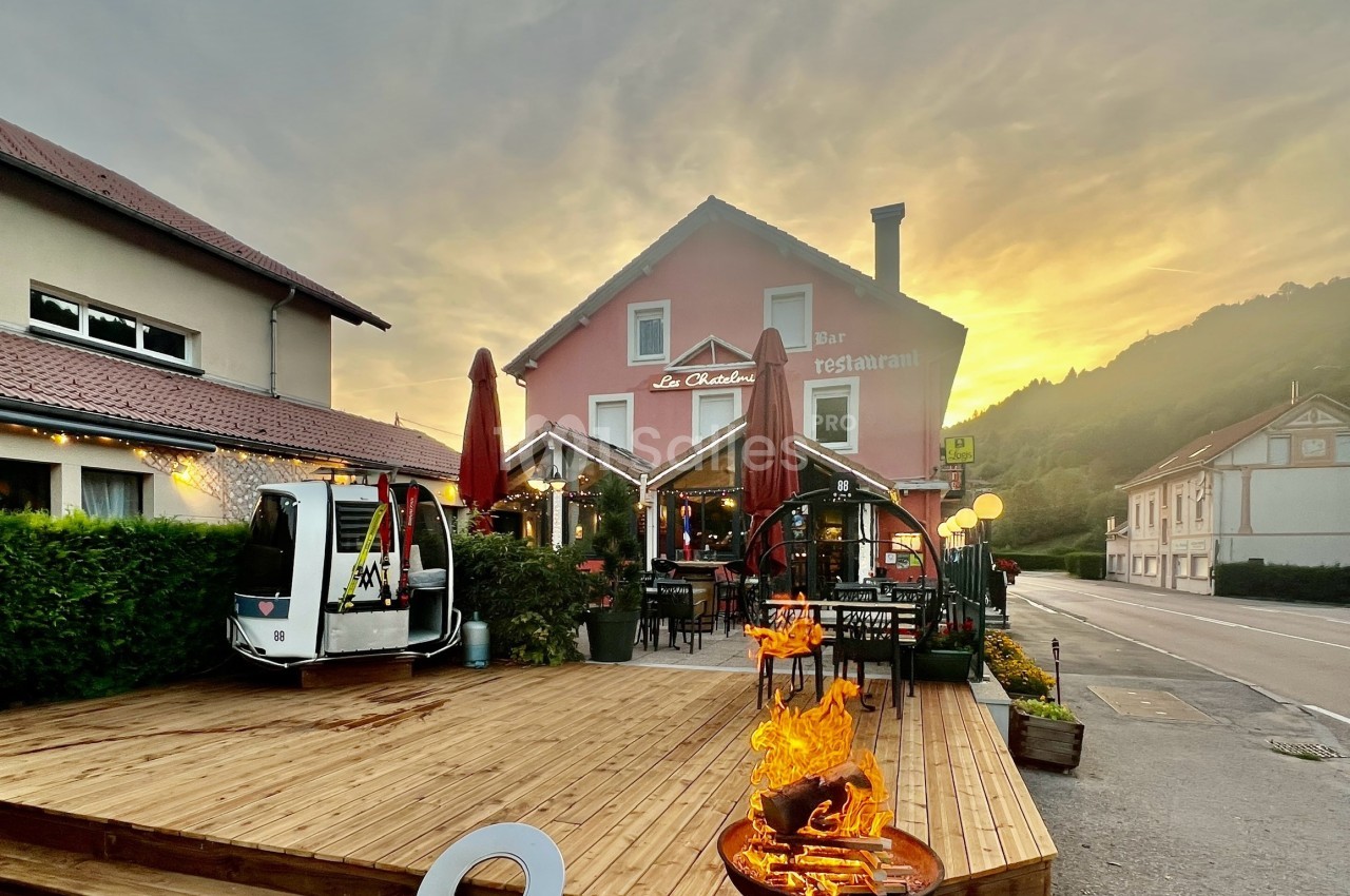 Terrasse en bois d'un restaurant avec brasero allumé, bâtiments environnants et coucher de soleil en arrière-plan.