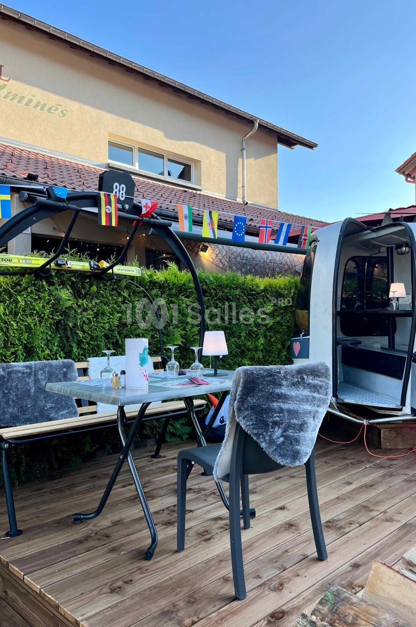 Terrasse en bois avec table dressée, chaises, télécabine aménagée et guirlande de drapeaux devant un bâtiment.