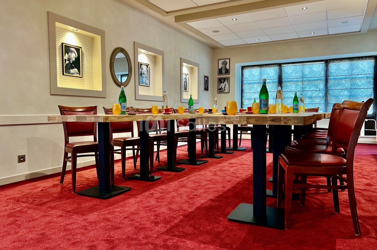 Salle de réunion avec tables alignées, chaises en bois, bouteilles d'eau et lumière naturelle filtrée par des stores.