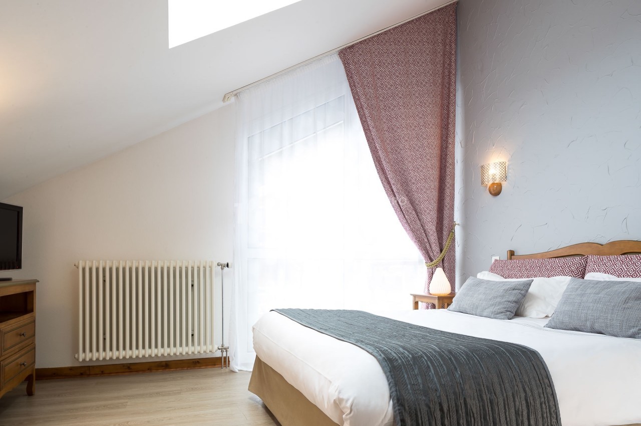 Chambre lumineuse avec lit double, draps blancs, plaid gris, rideaux rouges et mur gris, éclairée par une fenêtre.