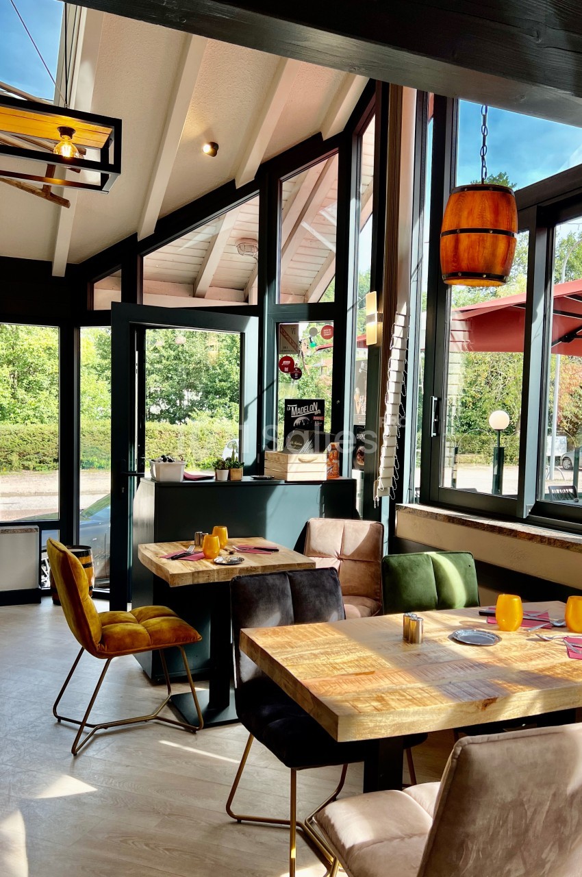 Salle de restaurant lumineuse avec grandes baies vitrées, tables en bois, chaises colorées et vue sur une terrasse…