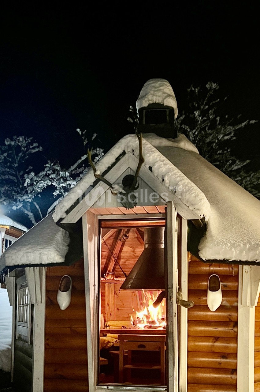 Cabane en bois enneigée avec un feu allumé à l'intérieur, vue de nuit dans un paysage hivernal.