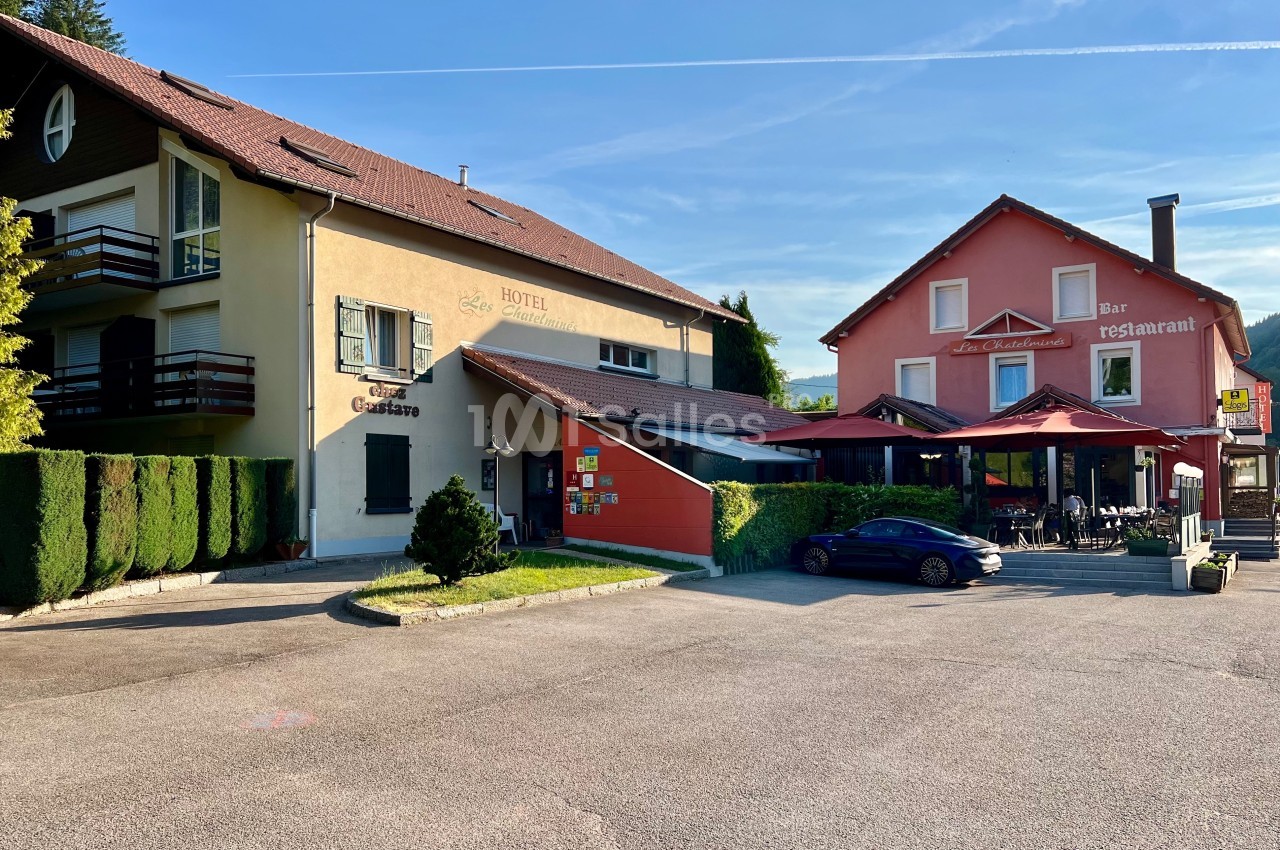 Façade d'un hôtel-restaurant avec terrasse, entouré de végétation et d'un parking sous un ciel dégagé.