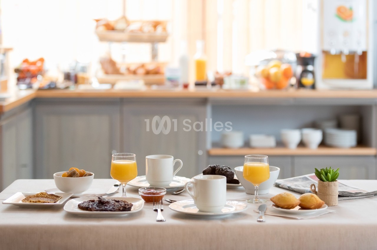 Petit-déjeuner servi sur une table avec viennoiseries, boissons chaudes, jus d'orange et confitures, en arrière-plan un…
