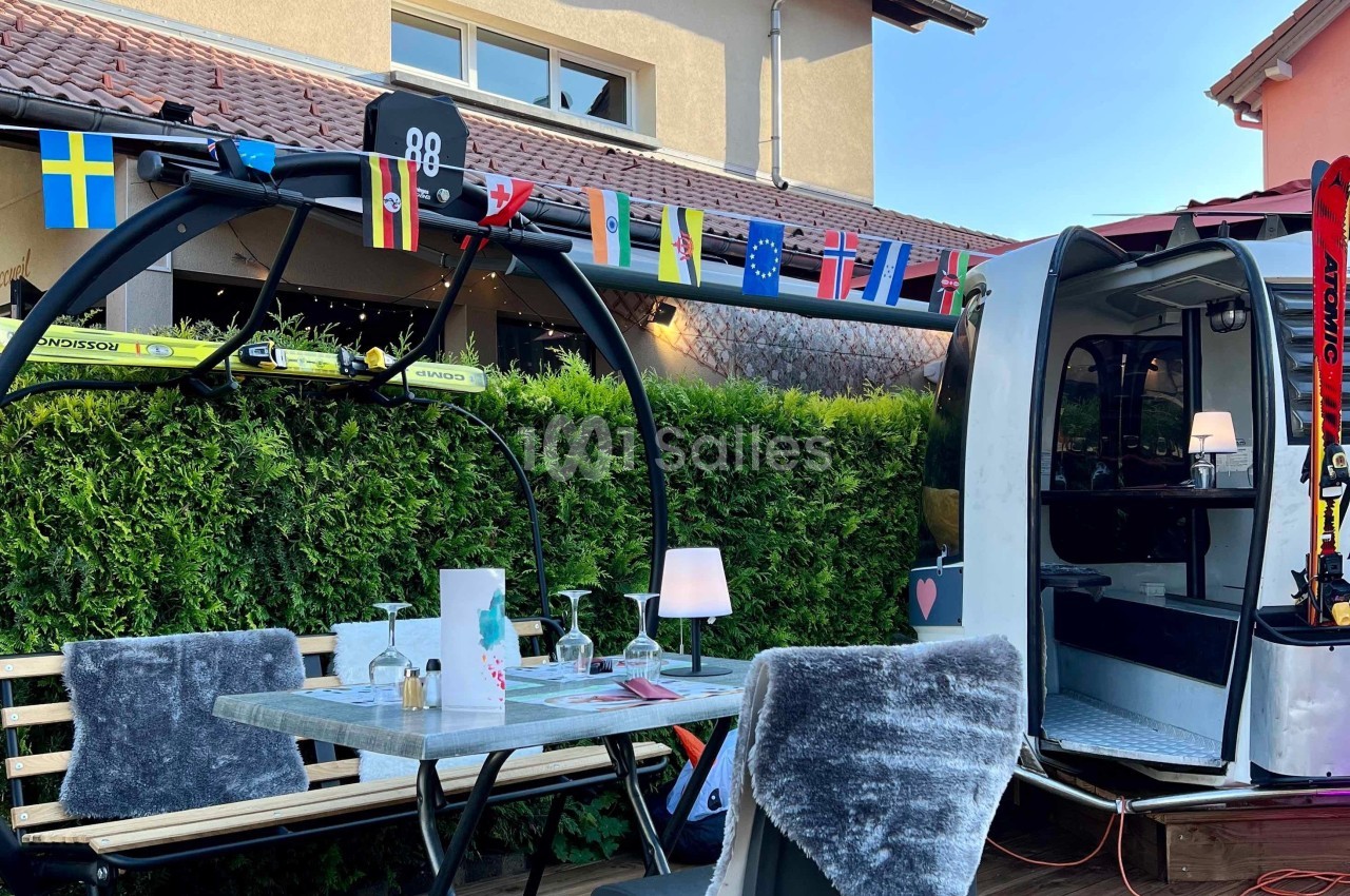 Table en extérieur avec des chaises couvertes de plaids, située près d'une cabine de télécabine décorée de drapeaux.